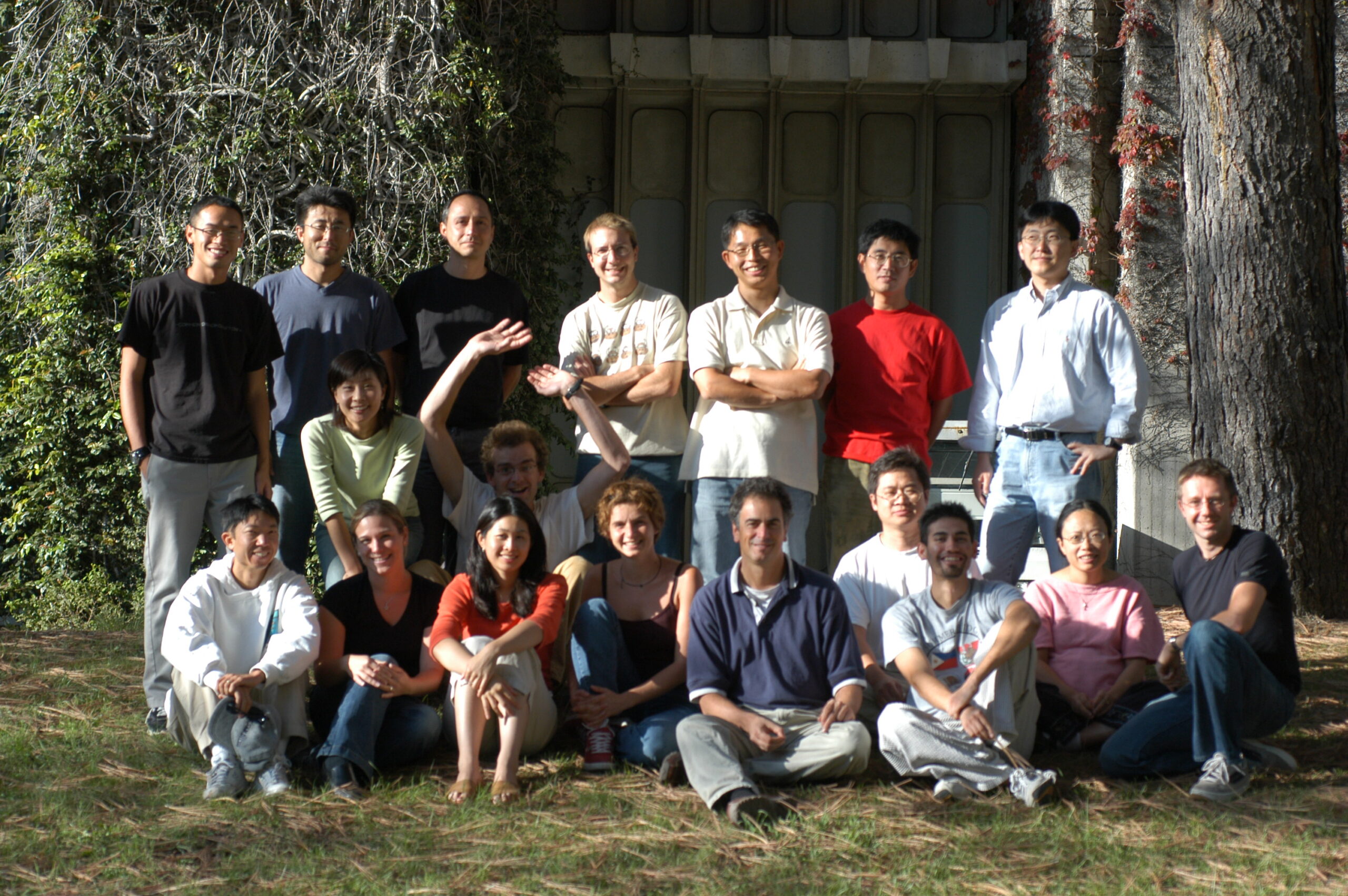 Schroeder lab members in two rows posing for a group photo. Some people sit on the grass in the front while others stand in the back. They smile at the camera with a concrete building behind them.