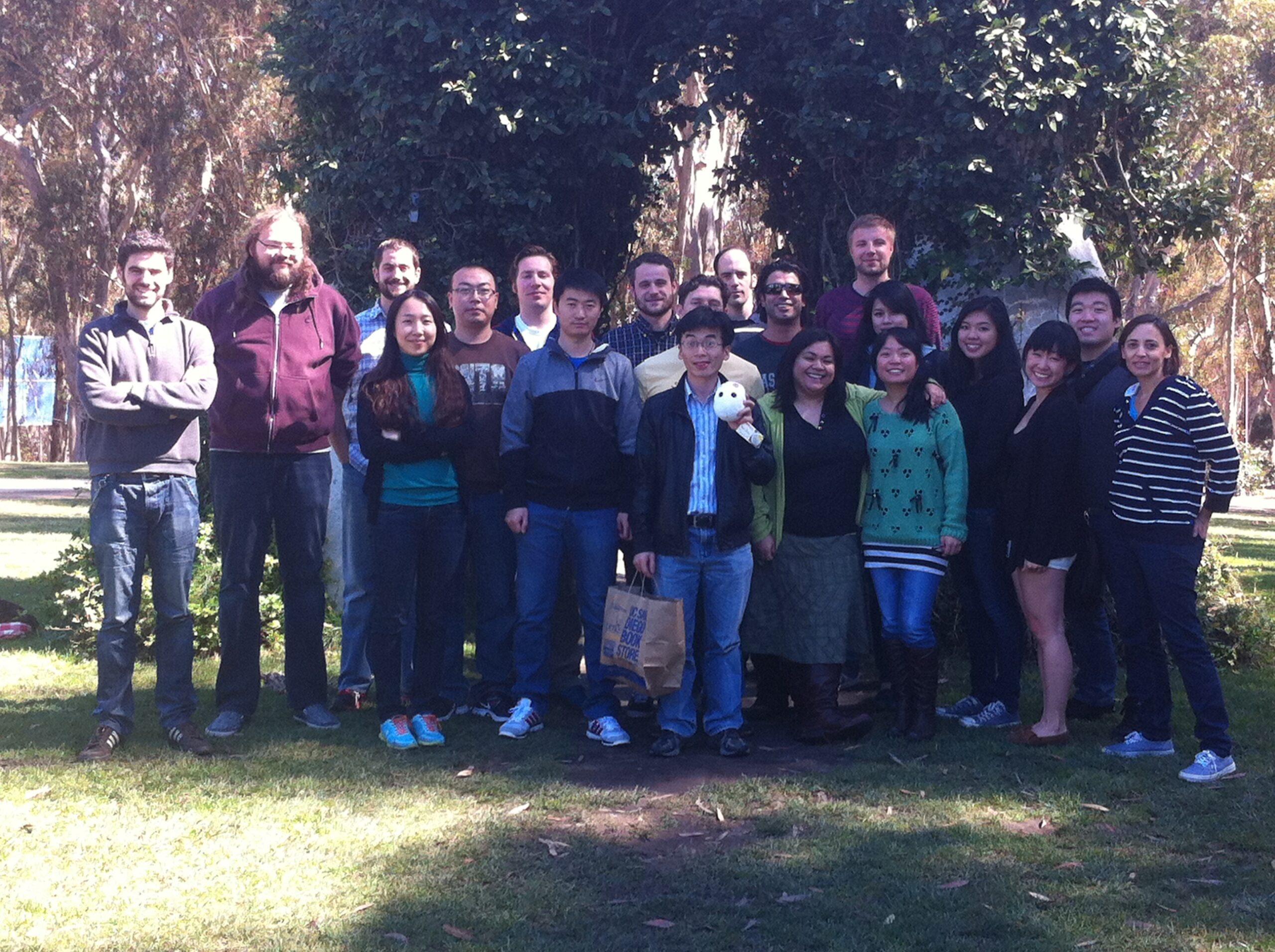 A group of 20 people stand in two rows in front of a green ivy arch. They all wear long sleeves and the man in the center holds a small egg-shaped stuffed animal as they all smile at the camera.