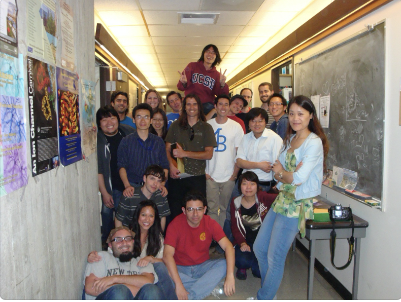 A group of people stand crammed together in a lab hallway. Some lean against the table while others sit on the floor. One person is sitting on two people's shoulders high above the rest making peace signs with their hands.