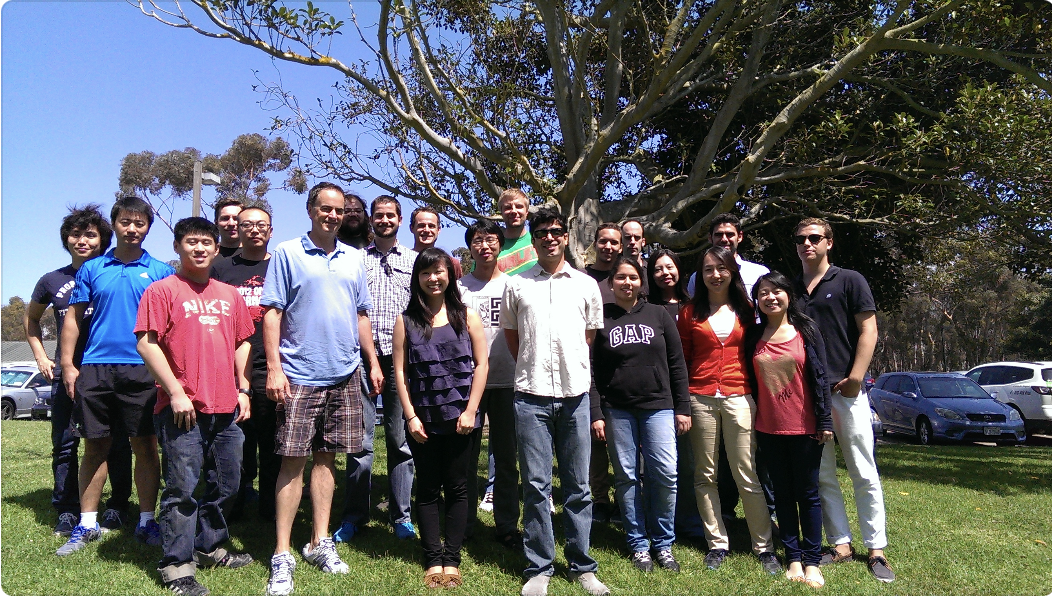 A group of 20 people stand in 3 rows in front of a large tree. They are smiling at the camera and most are wearing jeans or shorts with a t-shirt.